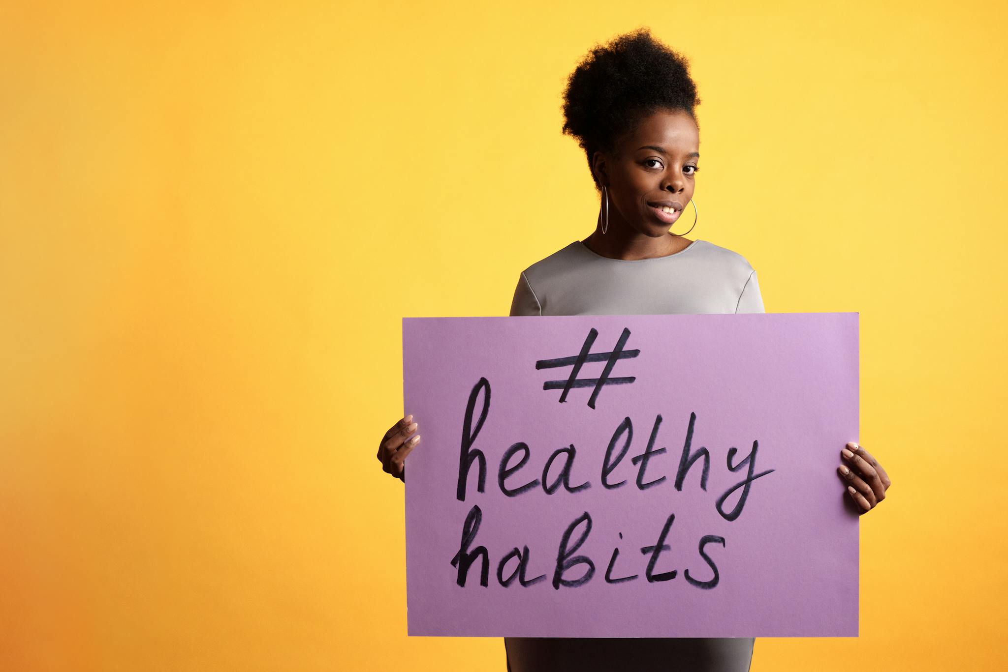 Portrait of a woman holding a healthy habits sign, set against a vibrant yellow background.